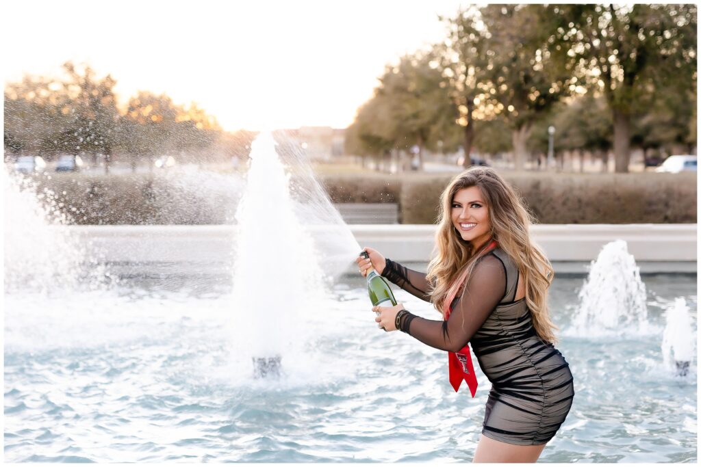 Woman in black sheer dress giving guns up while standing in a fountain and spraying champagne
