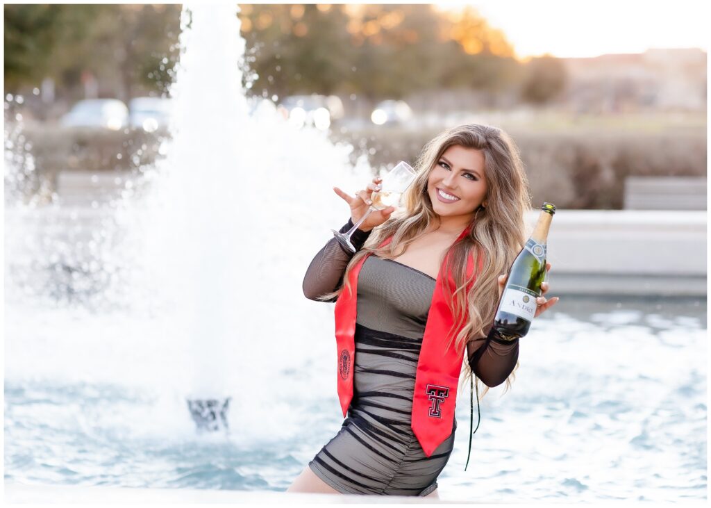 Woman in black sheer dress giving guns up while standing in a fountain and holding a champagne bottle