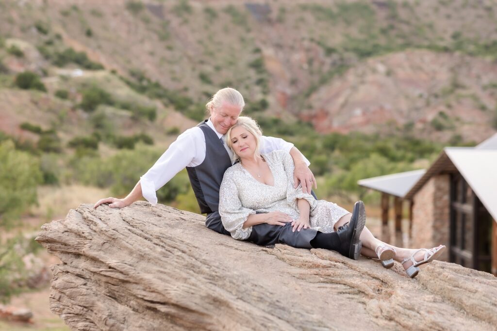 A bride and groom in wedding attire, sitting on a large rock in a scenic outdoor setting. The bride is wearing a silver sequin dress, while the groom is dressed in a suit. They are smiling and looking at each other, surrounded by nature.