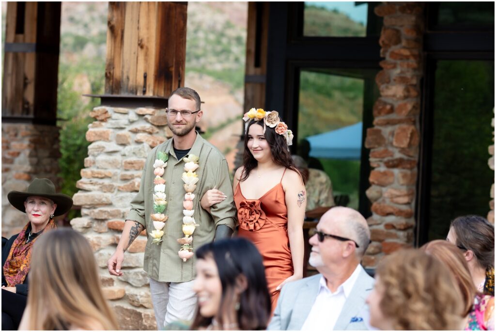Man escorting woman down the aisle with both wearing flowers in outdoor wedding.